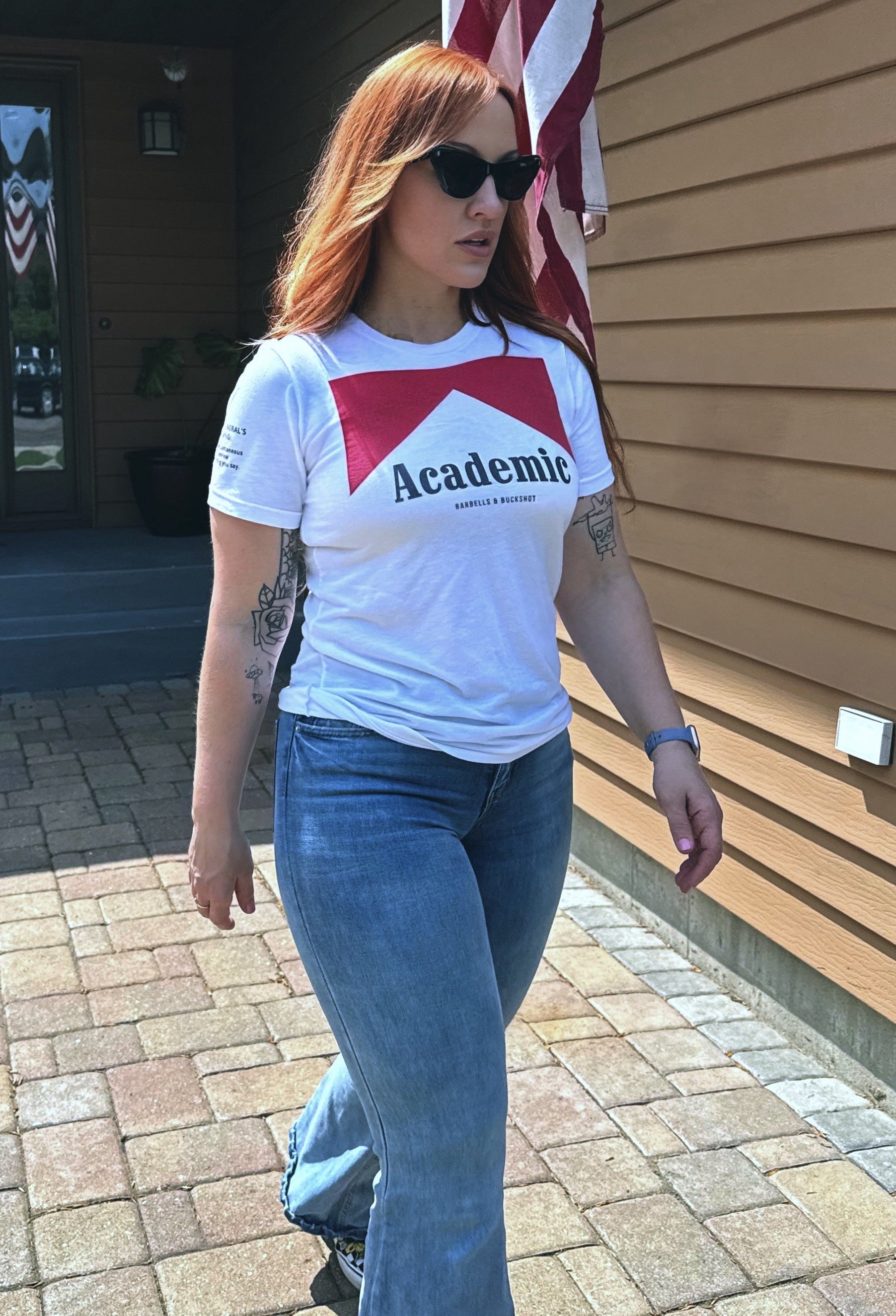 Person wearing a white t-shirt with red and blue design, standing outdoors near a wooden building and American flag.